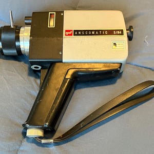 May include: A vintage black and silver GAF Anscomatic S/84 8mm film camera with a black leather strap. The camera has a built-in light meter and a zoom lens.