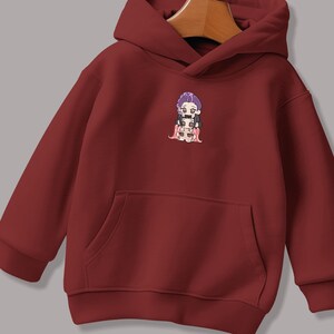 May include: A maroon hoodie with a kangaroo pocket and a hood. The hoodie features a small embroidered graphic of a character with purple hair. The graphic is located on the upper left chest area of the hoodie.