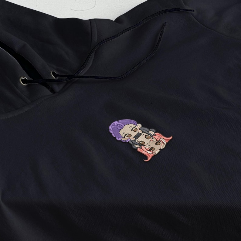 May include: Black hoodie with a drawstring hood. The hoodie features a small, embroidered patch of a cartoon character with purple hair and a pink cape on the front.