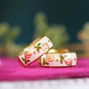 Może przedstawiać: Dwa złote pierścionki z białym emaliowanym paskiem. Każdy pierścionek ma kwiatowy wzór z różowymi i czerwonymi różami, zielonymi liśćmi i złotymi akcentami. Pierścionki są prezentowane na różowej powierzchni.