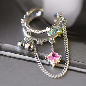 May include: Silver ear cuff with a double band design. The cuff features small, colorful gemstones and dangling charms, including a pink square gem and silver spheres, connected by delicate chains. The jewelry is set against a blurred background.