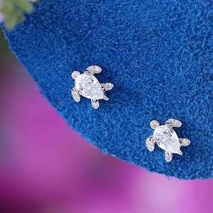 May include: A pair of silver-toned turtle stud earrings. Each earring features a pear-shaped, clear gemstone for the turtle's body, surrounded by smaller stones. The earrings are displayed on a textured, blue surface.