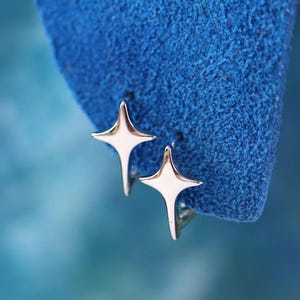 May include: A pair of silver star-shaped earrings. Each earring has a four-pointed star design with a smooth, reflective surface. The earrings are set against a textured blue background, creating a contrast in colour and texture. The earrings are small and delicate.