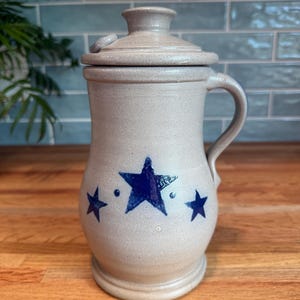 May include: A light gray stoneware crock with a matching lid and a single side handle. The exterior features a hand-painted design of three dark blue stars of varying sizes, with two small blue dots positioned between the central and side stars.