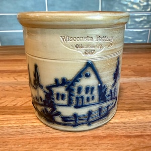 May include: A beige ceramic crock with a blue embossed landscape design featuring a house and trees. The text Wisconsin Pottery Columbus WI 1987 is stamped on the upper front. The vessel has a thick, rounded rim and a cylindrical body.