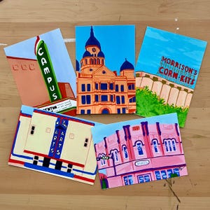 May include: Five colorful paintings of buildings in a city. The paintings are on white cardstock and feature a variety of architectural styles, including a courthouse, a movie theater, and a building with a sign that says "Morrison's Corn Kits".