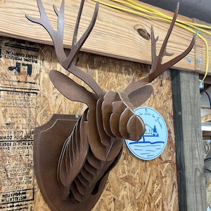 Puede incluir: Decoración de pared con cabeza de ciervo de madera marrón en capas, montada sobre una placa en forma de escudo. La cabeza de ciervo presenta astas detalladas y un diseño tridimensional. Un letrero azul y blanco con texto es visible a la derecha.