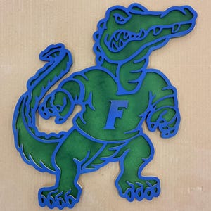 May include: A layered wooden cutout of a green alligator with blue outlines. The alligator is wearing a blue shirt with a blue "F" on it. The alligator is in a flexing pose.