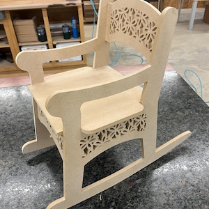 Peut inclure: Une chaise berçante en bois avec un motif floral découpé au dos et au bas. La chaise est en bois brut et a un design simple et classique.