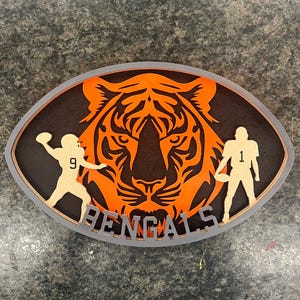 Könnte beinhalten: Ein Holzausschnitt eines Fußballs mit einem orangefarbenen Tigerkopf in der Mitte. Der Tiger hat schwarze Streifen und eine schwarze Nase. Die Worte "BENGALS" sind unter dem Tiger geschrieben. Zwei Fußballspieler stehen auf beiden Seiten des Tigers, einer mit der Nummer "9" und der andere mit der Nummer "1".