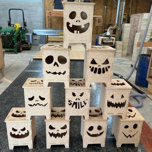 May include: A stack of nine wooden jack-o'-lantern candle holders with various facial expressions, including a smiling one that says "SMILEY".