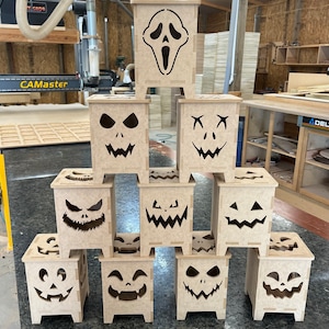 Puede incluir: Un conjunto de diez cajas de linterna de Halloween de madera con diferentes caras espeluznantes talladas en ellas. Las cajas están apiladas en forma de pirámide, con la caja superior que presenta una cara de fantasma gritando.