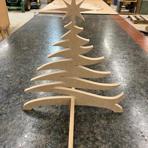 Può includere: Decorazione per albero di Natale in legno con una stella in cima. L'albero ha un design a strati unico con rami curvi. La decorazione è realizzata in legno marrone chiaro e si erge su una base di legno.