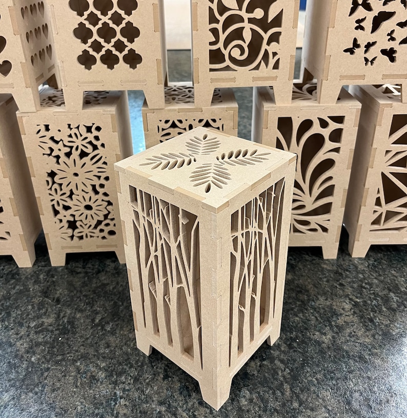 May include: A set of wooden candle holders with intricate laser cut designs. The designs include floral patterns, geometric shapes, and a tree branch pattern.