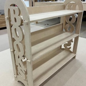 May include: A light-colored wooden bookshelf with three shelves. The word "BOOKS" is cut out of the side supports. The shelves are rectangular and the overall design is simple and functional. The bookshelf is made of unfinished wood.
