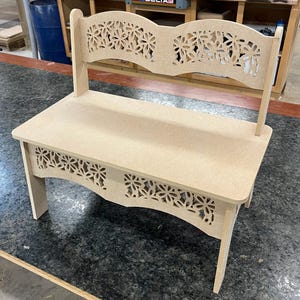 May include: A light-colored wooden bench with a seat, back, and legs. The back and the apron of the bench feature a floral cut-out design. The bench is made of unfinished wood.