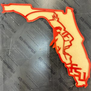 May include: A layered wooden cutout in the shape of Florida, with a red outline and a light tan interior. The image features a Seminole figure and the letters "FSU" in red. The cutout is likely a decorative item or a piece of art.