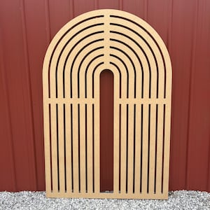 Puede incluir: Un arco de madera con un diseño de corte de múltiples arcos de arcoíris. El arco está hecho de madera marrón claro y está de pie sobre una superficie de grava.