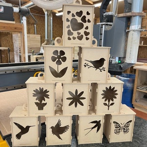 Puede incluir: Un conjunto de nueve portavelas de madera con varios diseños recortados, incluyendo corazones, flores, pájaros y mariposas. Los portavelas están hechos de madera sin terminar y están listos para ser pintados o teñidos.