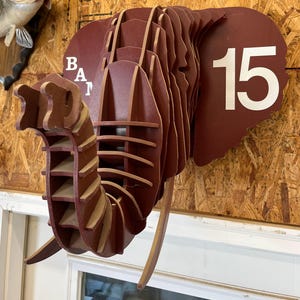 May include: A layered, three-dimensional elephant head sculpture in shades of brown and tan. The sculpture features the number "15" in white and the letters "BAT" in white. The elephant's trunk is detailed with horizontal lines.
