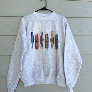 May include: A heather gray sweatshirt featuring a series of colorful, stylized, vertical designs. The designs are in shades of blue, red, brown, and gold. The sweatshirt has a crew neck and long sleeves.