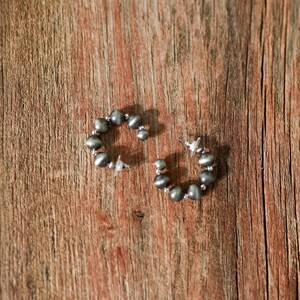 Puede incluir: Un par de pendientes de aro de plata con una fila de cuentas de color gris oscuro. Los pendientes están sobre una superficie de madera.
