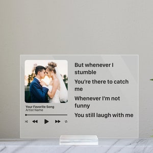 Puede incluir: Un marco de fotos de acrílico transparente con un diseño de reproductor de música en blanco y negro. El marco presenta una foto de una pareja en un entorno de boda. El texto en el marco dice "But whenever I stumble You're there to catch me Whenever I'm not funny You still laugh with me".