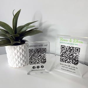 May include: Two clear acrylic signs with QR codes and text. The top sign reads "Renew & Bloom Health & Wellness Spa". The bottom sign reads "Connect with us!" and includes social media icons and the text "@feellikehomeshop".