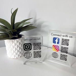 以下が含まれることがあります： 黒い文字とQRコードが書かれた透明なアクリル製の看板が2つ。最初の看板には「Let's get social!」と書かれており、Instagram、Facebook、地球儀のアイコンが描かれている。2番目の看板には「Connect with us」と書かれており、Facebook、Instagram、地球儀のアイコンが描かれている。両方の看板には「scentedserenade.com」というウェブサイトが印刷されている。