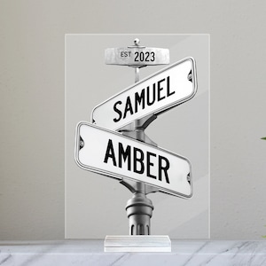 May include: A clear acrylic sign with a silver metal base featuring two street signs. The top sign reads "EST. 2023" and the bottom sign reads "SAMUEL" and "AMBER".