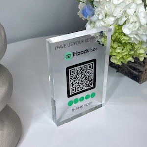 May include: A clear acrylic sign with the text "Leave us your review on TripAdvisor" and a TripAdvisor logo. A black and white QR code is below the text, followed by five green dots and the text "Thank you!"