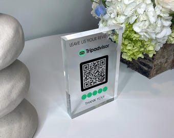 May include: A clear acrylic sign with the text "Leave us your review on TripAdvisor" and a TripAdvisor logo. A black and white QR code is below the text, followed by five green dots and the text "Thank you!"