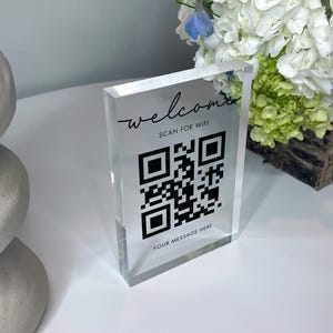 May include: A clear acrylic sign with the word "welcome" in script, "SCAN FOR WIFI", a QR code, and "YOUR MESSAGE HERE" in black. The sign is rectangular and stands upright. A stack of white stones and a floral arrangement are in the background.