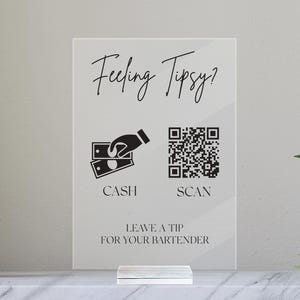 May include: A white sign with black text that reads "Feeling Tipsy?" with a cash icon and a QR code icon below. The text below the icons reads "Leave a tip for your bartender".