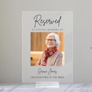 May include: A clear acrylic sign with the word "Reserved" in elegant script. Below the text is a photo of a woman wearing glasses and a red scarf. The sign reads "In Loving Memory of Grace Jones, Grandmother of the Bride."