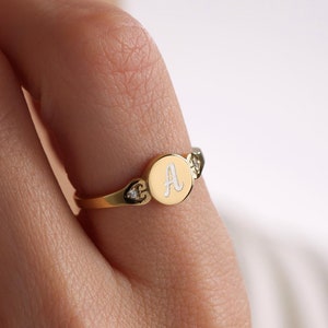 May include: A gold ring with a round, white enamel center featuring the letter "A". The ring is accented with two small, clear stones on either side of the center.