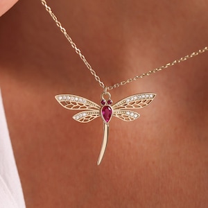 May include: A gold necklace with a dragonfly pendant. The dragonfly has a red gemstone in its body and clear gemstones on its wings.
