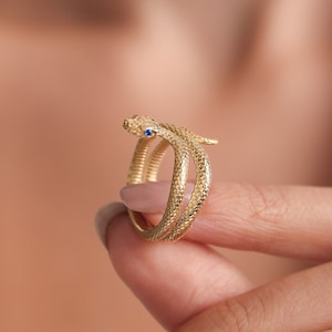 May include: A gold snake ring with blue eyes. The ring is coiled around a finger and the snake's head is facing forward.