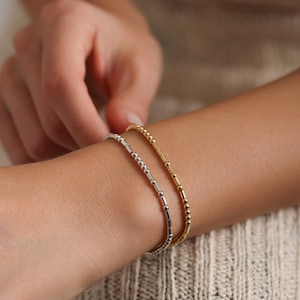 May include: Two beaded bracelets on a person's wrist. The top bracelet is gold and the bottom bracelet is silver.
