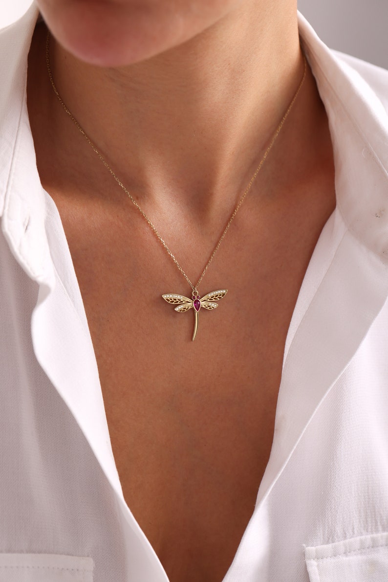 May include: A gold necklace with a dragonfly pendant featuring a red gemstone. The dragonfly has intricate details and a delicate design.