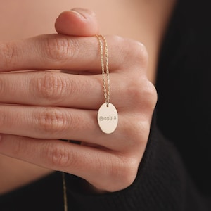 May include: A gold-colored necklace with a delicate chain and an oval pendant. The pendant is engraved with the name "Sophia" in an elegant script. The necklace is held up against a dark background.