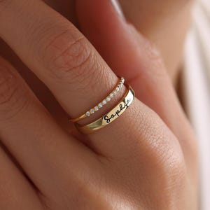 Może przedstawiać: Złoty pierścionek z cienkim paskiem błyszczących kamieni i szerszym paskiem grawerowanym imieniem "Sophia".