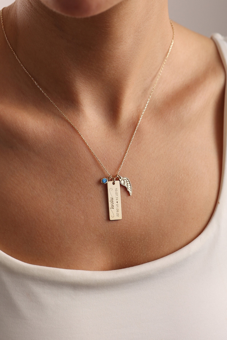 Puede incluir: Collar de oro con una peque&ntilde;a piedra preciosa azul, un dije de ala y una etiqueta rectangular grabada con "mom" y las fechas "10.07.74" y "01.02.22".