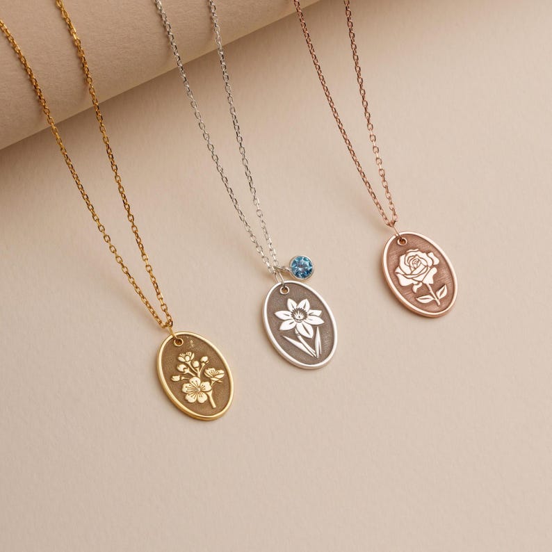May include: Three necklaces with oval pendants, each featuring a different floral design. One is gold-toned with a flower cluster, another is silver with a daffodil, and the third is rose gold with a rose. A blue gemstone accents the silver necklace.