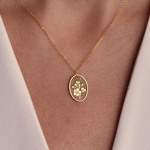May include: A gold-colored necklace with an oval pendant featuring a floral design. The pendant hangs from a delicate chain. The floral design is detailed and appears to be etched into the pendant's surface. The necklace is worn against a white background.
