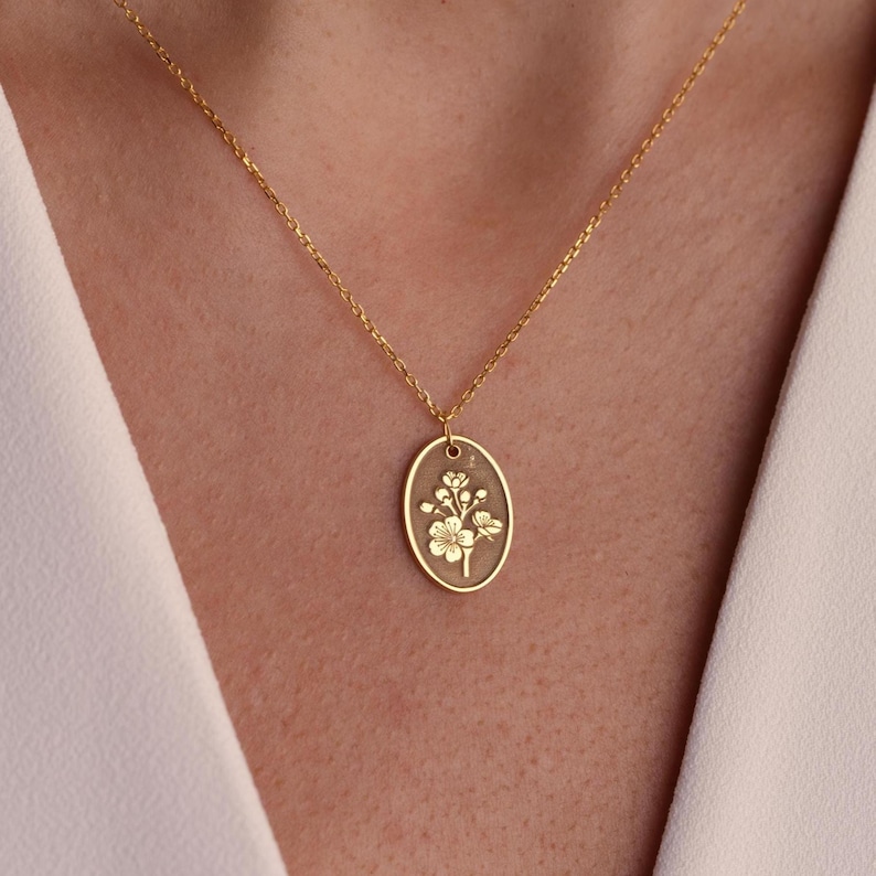 May include: A gold-colored necklace with an oval pendant featuring a floral design. The pendant hangs from a delicate chain. The floral design is detailed and appears to be etched into the pendant's surface. The necklace is worn against a white background.