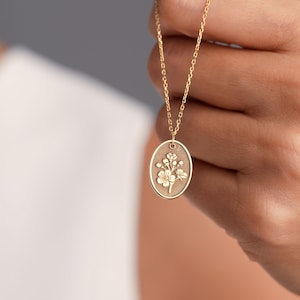 May include: Gold-colored necklace featuring an oval pendant with a floral design. The pendant hangs from a delicate chain. The necklace is held up against a neutral background, showcasing the jewelry's details and craftsmanship.