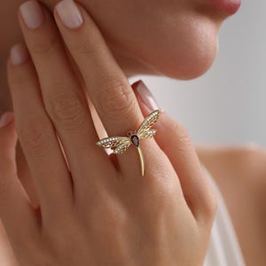 May include: A gold dragonfly ring with a red gemstone and clear accents. The ring is worn on a finger.