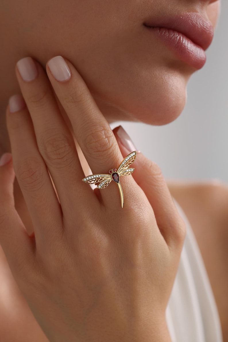 May include: A gold dragonfly ring with a red gemstone and clear accents. The ring is worn on a finger.