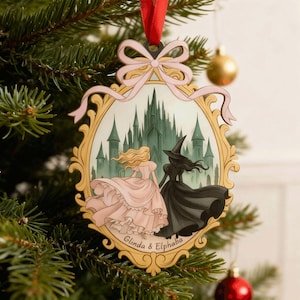 Puede incluir: Adorno navideño de madera con una ilustración enmarcada de dos mujeres con vestidos, uno rosa y otro negro, frente a un castillo. El adorno tiene un marco dorado, un lazo rosa y una cinta roja para colgar. El texto "Glinda & Elphaba" está en la parte inferior.